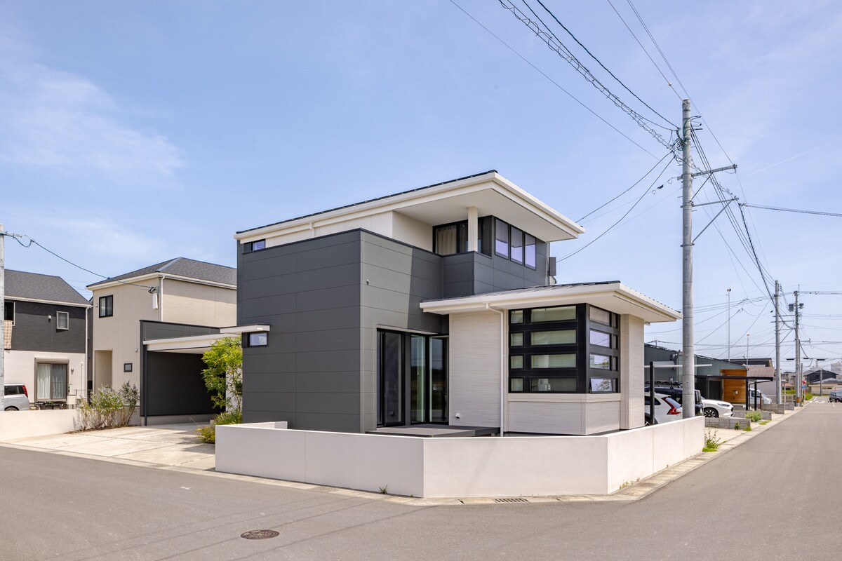 A modern two-story house features a combination of dark and light exterior materials, showcasing large windows that allow natural light to enter. A minimalist front yard is complemented by a clean driveway and nearby utility poles, contributing to the property's contemporary appeal.