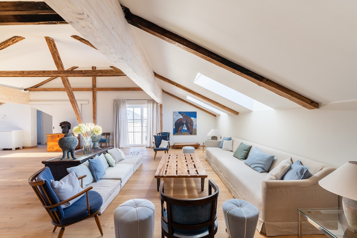 A spacious living area features tall ceilings with exposed wooden beams and skylights that allow natural light to fill the room. Elegant seating includes a mix of sofas and armchairs positioned around a long wooden table. A decorative piece of art is displayed on the wall.