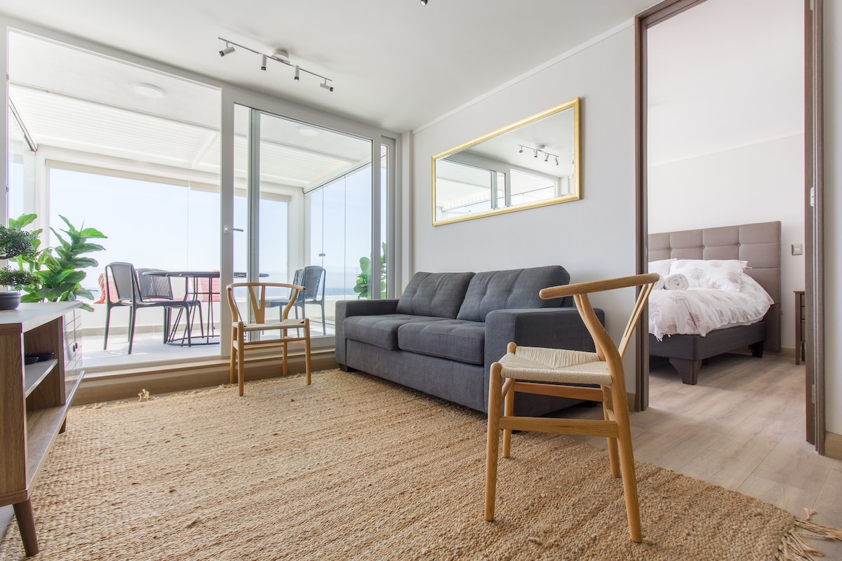 A living area features a comfortable gray sofa and two wooden chairs, set on a natural woven rug. Large glass doors open to a terrace with outdoor seating, allowing ample natural light to fill the space. A framed mirror reflects the interior design.