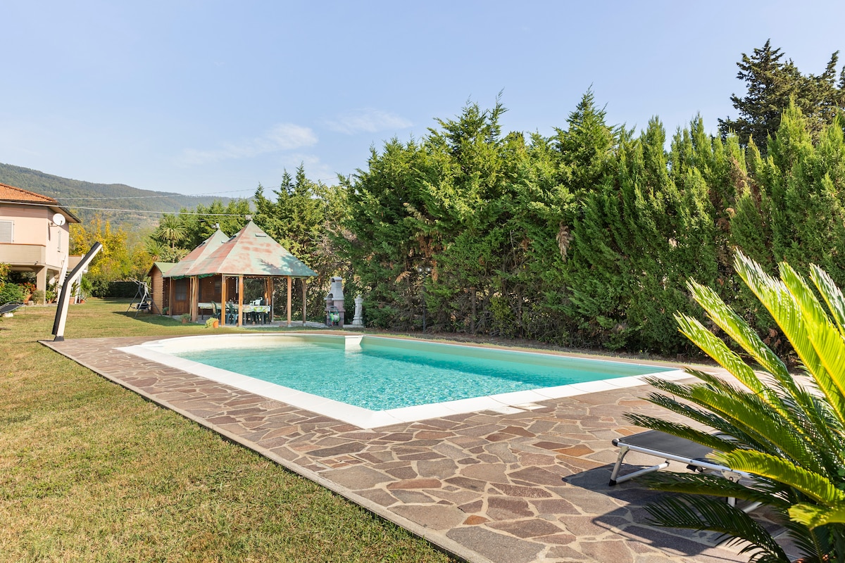 A spacious outdoor area features a salt-water pool surrounded by landscaped greenery. A gazebo with seating is positioned nearby, offering shade and a place to relax. Sun loungers are arranged along the poolside, providing options for sunbathing in a serene environment.