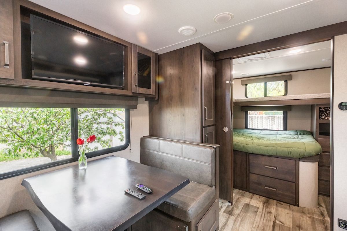 An indoor view of a modern camper features a dining booth with plush seating, accompanied by a small table. A television is mounted on a wall above, while a cozy sleeping area is visible through a partition, showcasing a green-covered bed. A vase with flowers is placed on the table.