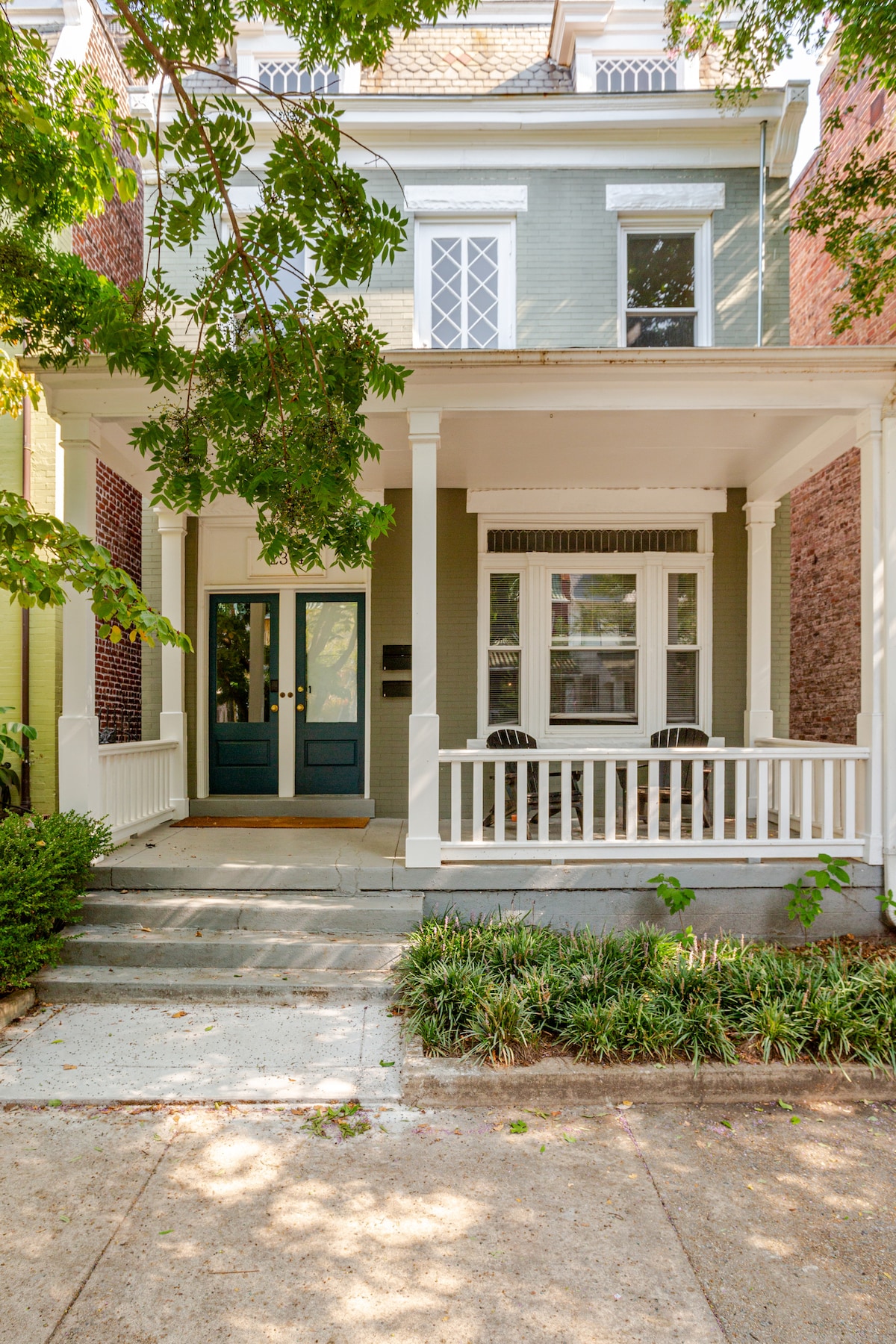 The exterior of the two-story building features a green facade with white trim and a welcoming porch. Two rocking chairs are situated on the porch, surrounded by lush greenery and a well-maintained pathway leading to the entrance.