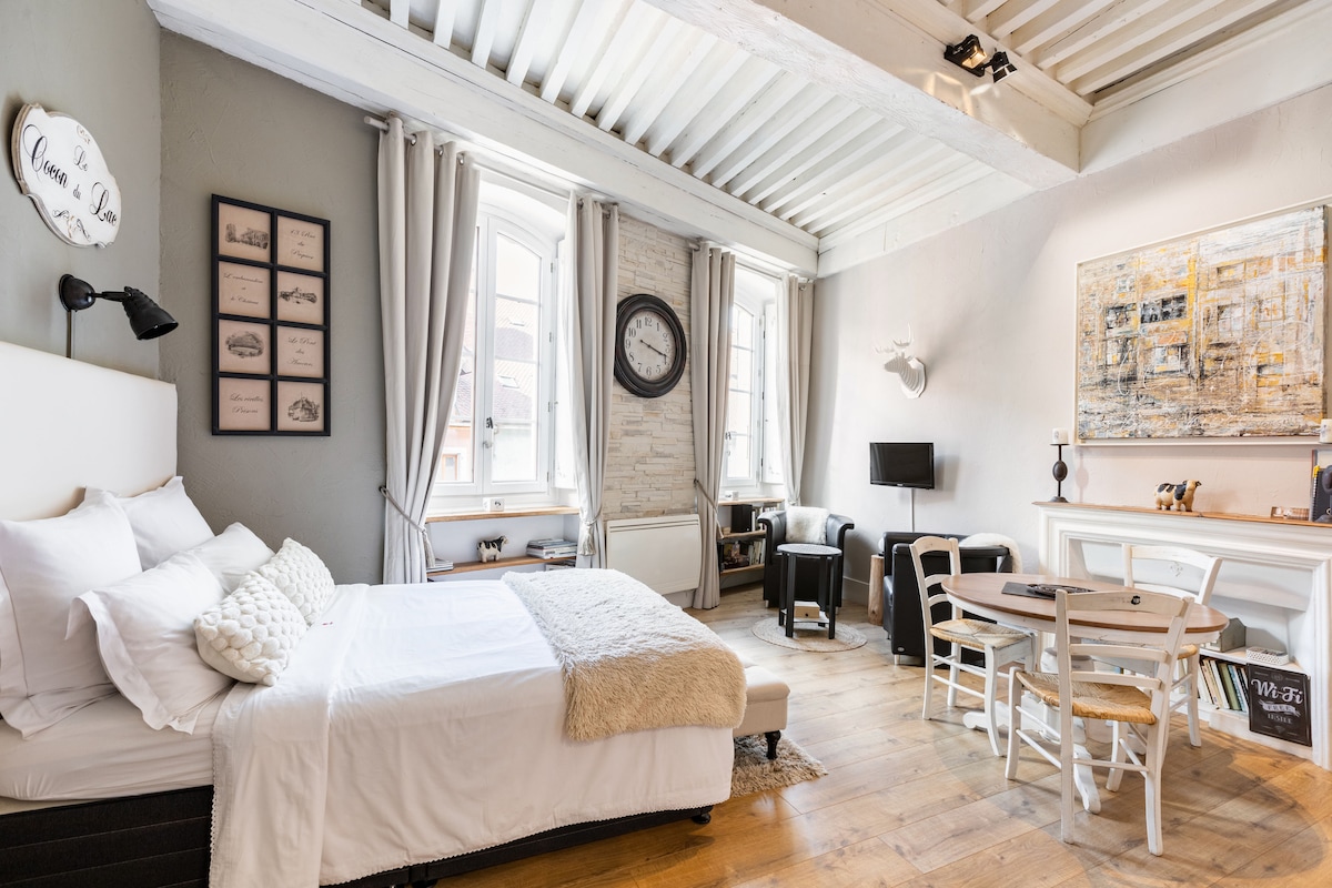 A bright and airy room features a spacious bed with soft linens. Natural light enters through large windows, framed by light curtains. The seating area includes a cozy chair and a small dining table. Artistic decor enhances the elegant yet cozy ambiance of the space.