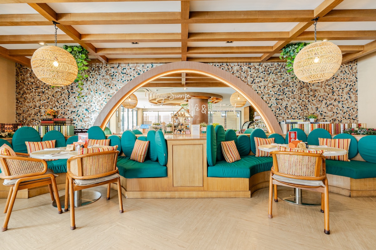 A cozy dining area features brightly colored seating with turquoise cushions surrounding a wooden table. Natural light streams in, highlighting the unique circular archway and decorative wall with a mosaic design. Rattan pendant lights provide a warm glow overhead.