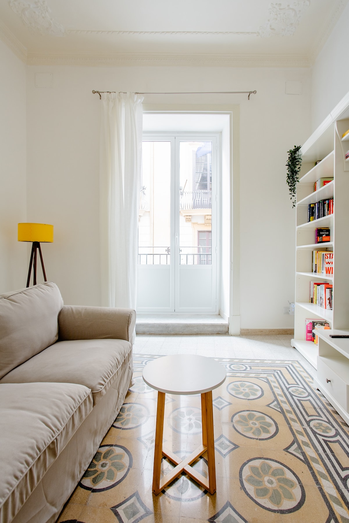 A light-filled living area features a beige sofa and a small round table, with pale curtains gently framing a set of double doors. A bookcase filled with books stands against one wall, and decorative tiles on the floor add a unique touch.