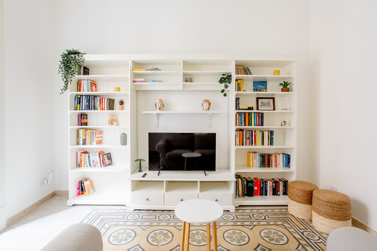 A bright living area is highlighted by a white bookshelf filled with a variety of books. A sleek television is mounted above a low cabinet. Two woven stools sit beside a small coffee table, while decorative items and plants add a touch of charm.