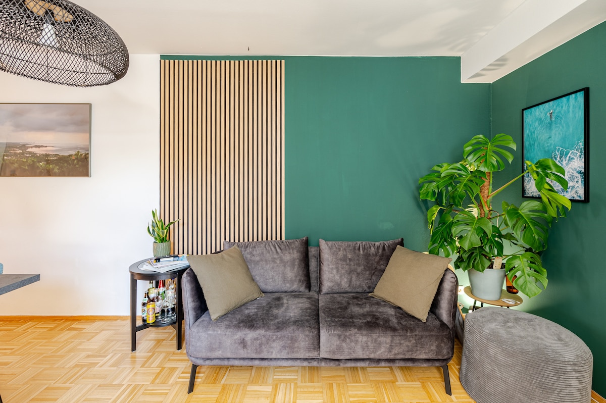 A spacious living area features a gray sofa adorned with soft cushions, positioned against a green wall. A round side table holds various items, and a large plant flourishes nearby. Natural light illuminates the space, creating a relaxed environment with modern decor.