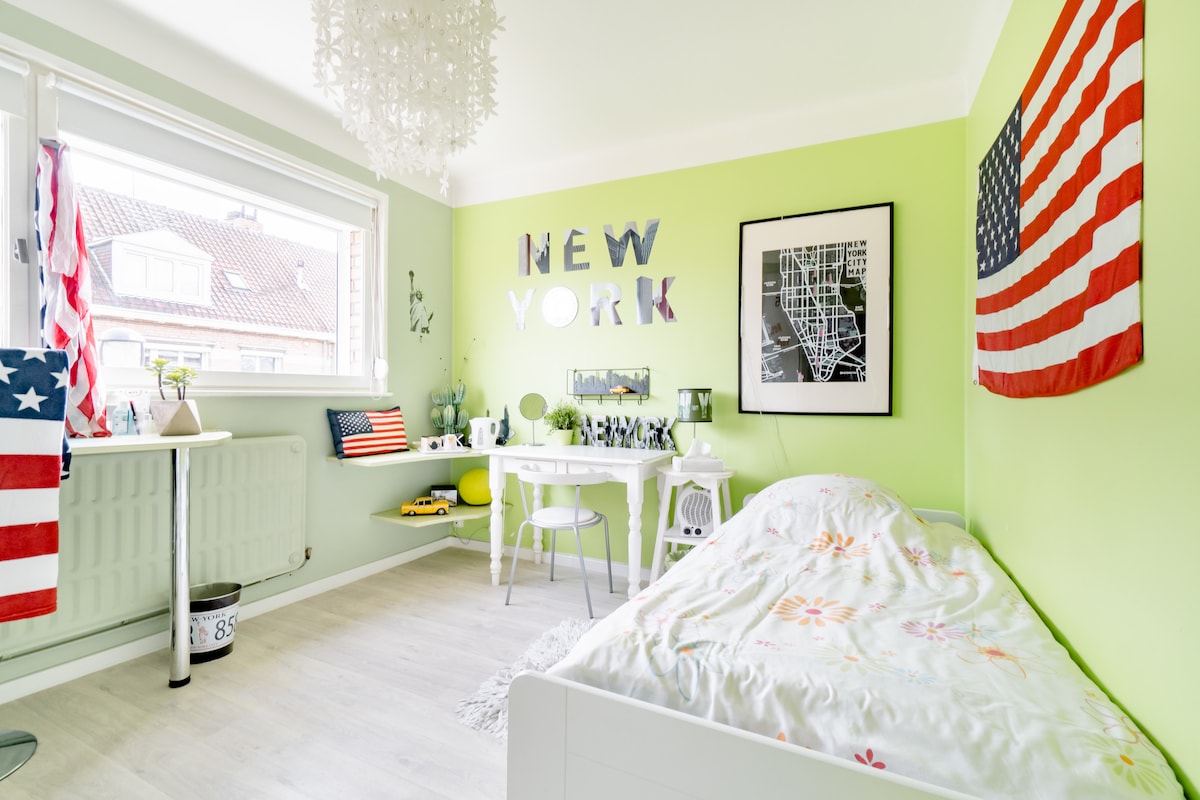 A bright room features green walls and natural light filtering in through the window. A bed with a floral bedspread is positioned against one wall, while a small table and chairs provide a workspace. American flags add a festive touch to the decor.