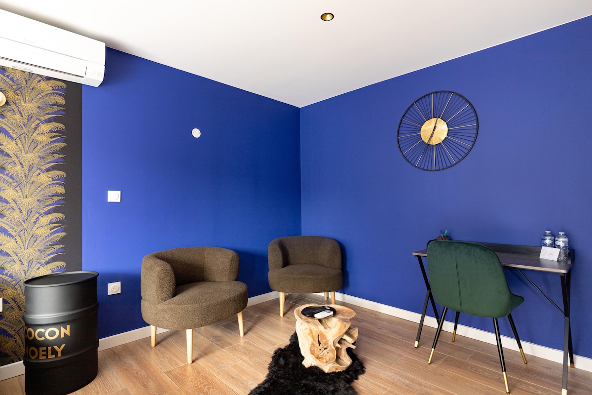 A vibrant blue wall serves as a backdrop for this cozy seating area, featuring two comfortable armchairs and a wooden accent table. A small desk with a green chair is provided for work needs. A decorative clock adds a touch of style to the space.