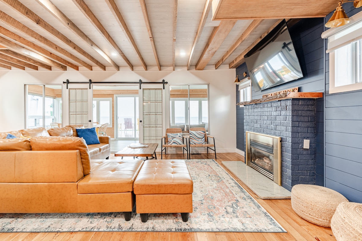 A spacious living area showcases a comfortable leather sectional sofa with an ottoman. A flat-screen television is mounted above a modern fireplace. Mirrored panels enhance the natural light, and sliding glass doors provide access to the outdoor area. A decorative area rug adds warmth to the space.