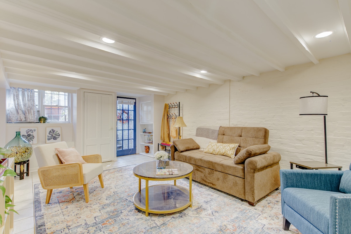 A spacious living area features a cozy brown sleeper sofa and a light armchair. A round coffee table sits in the center, with decorative items on display. Large windows provide natural light, and soft white beams span the ceiling, enhancing the room's inviting feel.