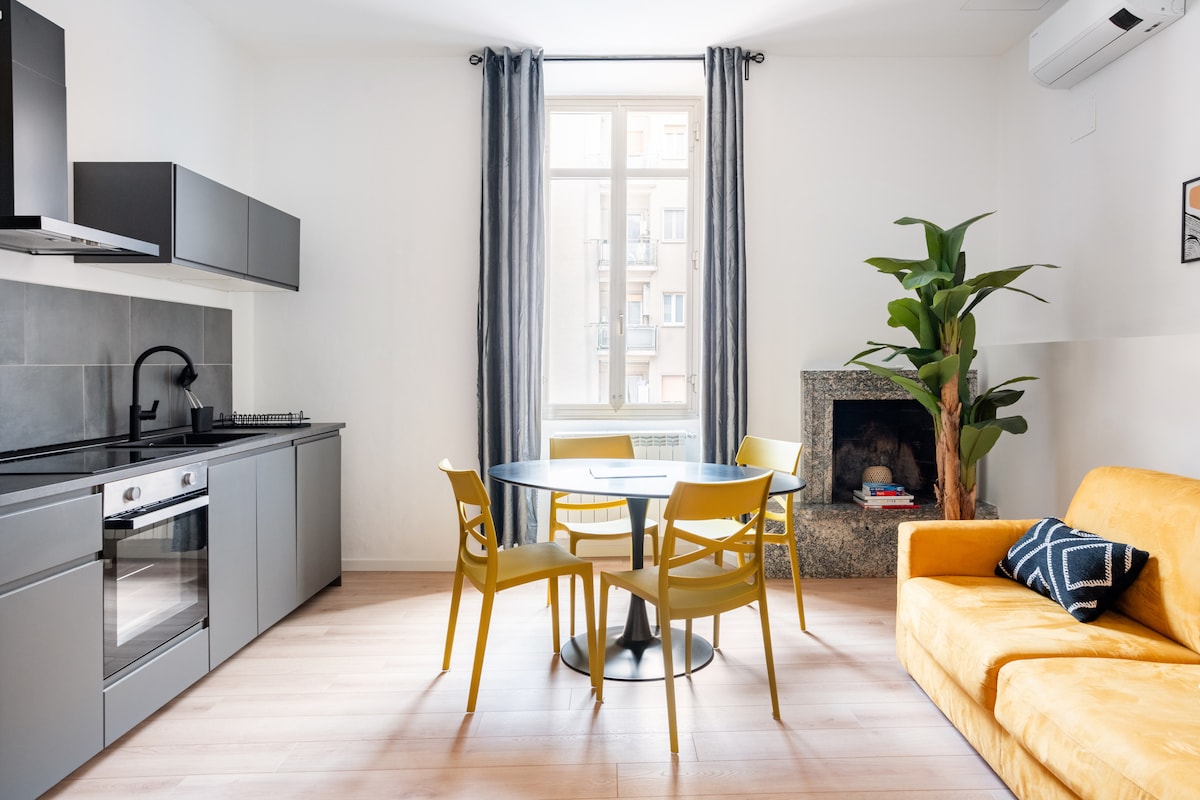An open-plan kitchen and living area features modern cabinetry and a gleaming countertop. A round dining table is surrounded by four yellow chairs. A plush sofa sits nearby, complemented by a potted plant and a window allowing natural light to illuminate the space.