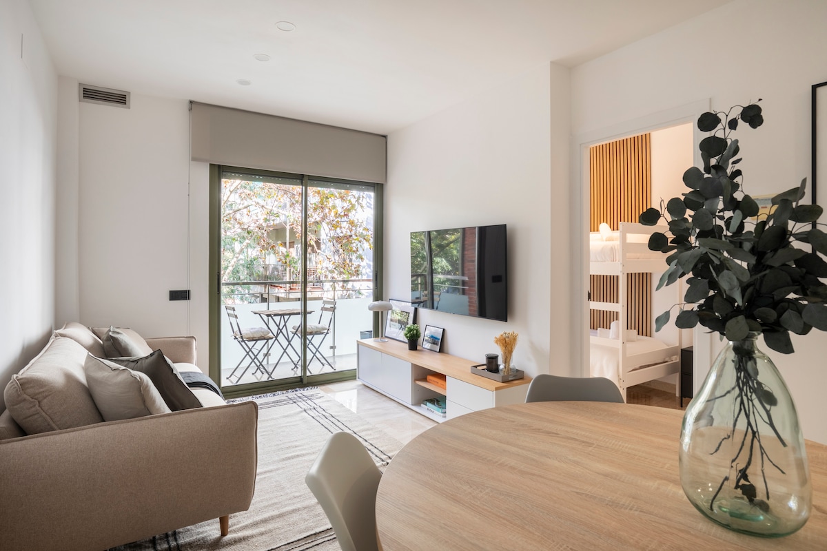 A light-filled living area features a comfortable sofa and a flat-screen TV mounted on a white cabinet. A small balcony is accessible through sliding glass doors. A dining table with a clear vase and greenery adds a touch of nature to the space.