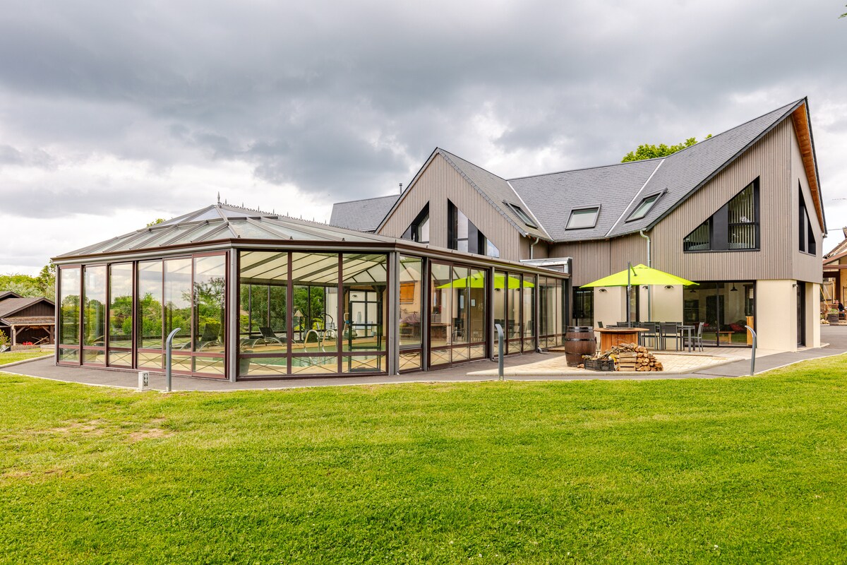 An architecturally designed home features a large glass sunroom that invites natural light. The structure is complemented by green outdoor umbrellas and is surrounded by well-maintained grass. The modern design incorporates an array of geometric shapes, offering a harmonious blend with the landscape.