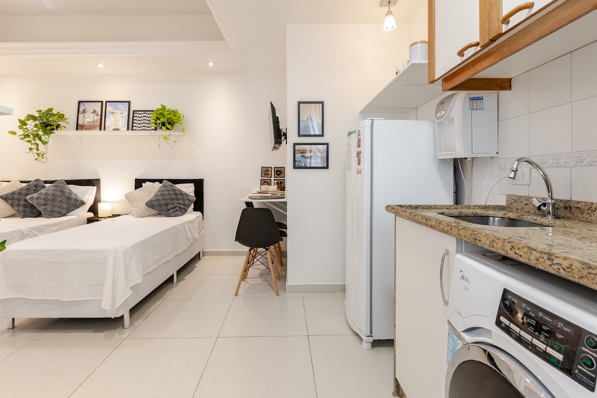 A compact studio apartment features two beds with neutral bedding positioned against a white wall. A small kitchenette with granite countertops and a washer is visible. Wall shelves display green plants and framed photos, while a mounted TV adds to the functionality of the space.