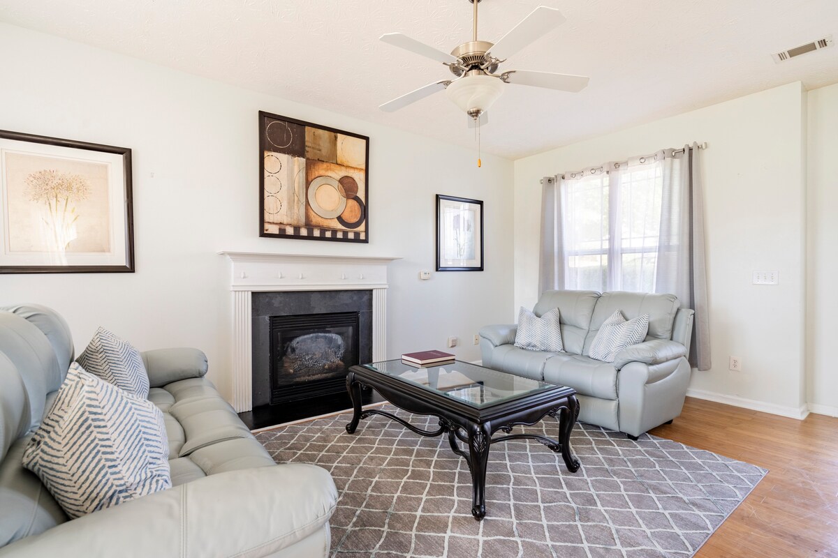 A light-filled living room is furnished with a pair of comfortable gray sofas and a coffee table. A decorative wall art piece is displayed above a fireplace, while a ceiling fan provides air circulation. Natural light filters through the sheer curtains, adding to the uplifting ambiance.