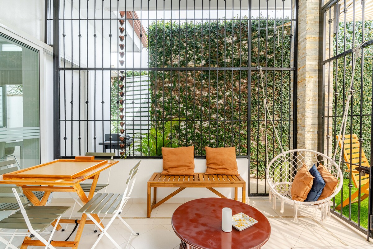 A spacious patio area is enclosed by a decorative metal lattice. Wooden furniture, including a circular table and chairs, is complemented by orange cushions. A hammock and additional seating are positioned against a backdrop of vibrant green foliage.