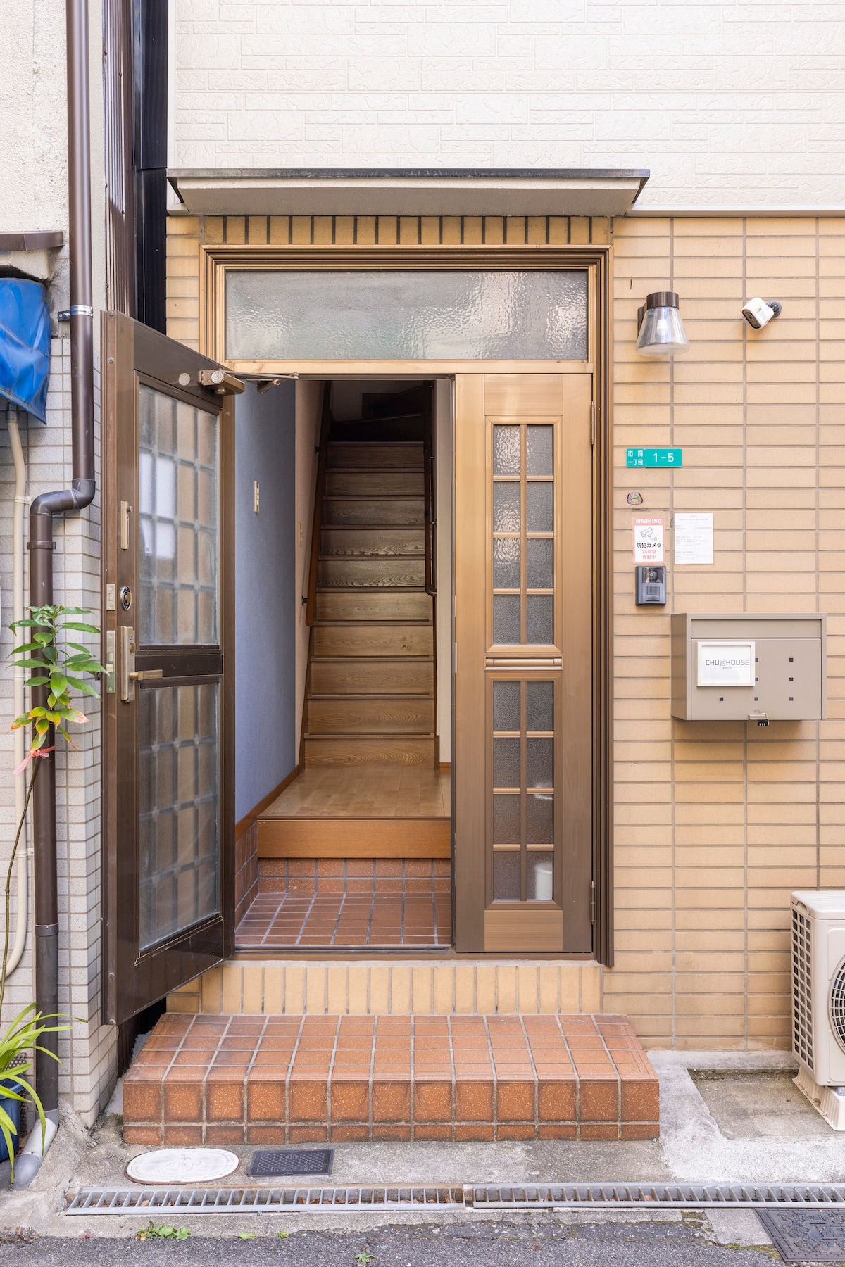 The entrance features a wooden door with glass panes, revealing a staircase leading to the upper floors. The entryway has tiled flooring, and a mailbox is mounted on the wall beside the door. The surrounding walls are made of light-colored brick.