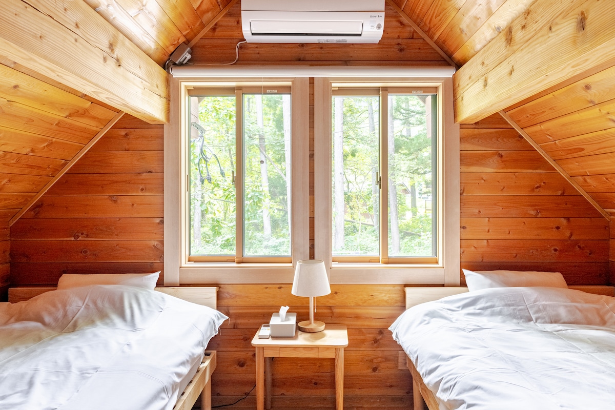 A cozy sleeping area features two spacious beds placed against a wooden wall. Natural light fills the room through large windows, while a small bedside table with a lamp stands in between. An air conditioning unit is mounted above the windows.