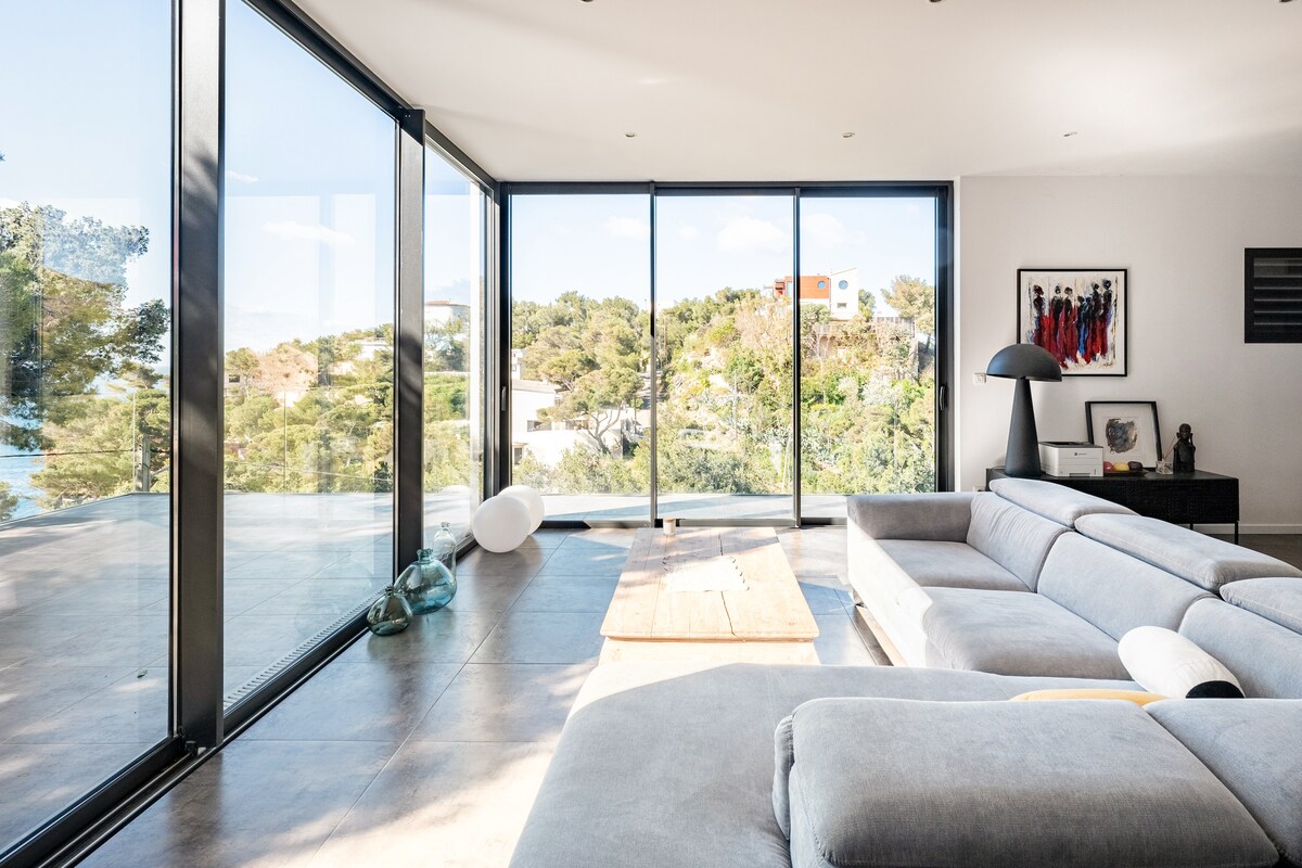 A spacious living area is defined by large glass walls that provide abundant natural light and a view of the surrounding landscape. A modern grey sofa is positioned adjacent to a low wooden coffee table, while artwork adorns one wall, adding character to the room.