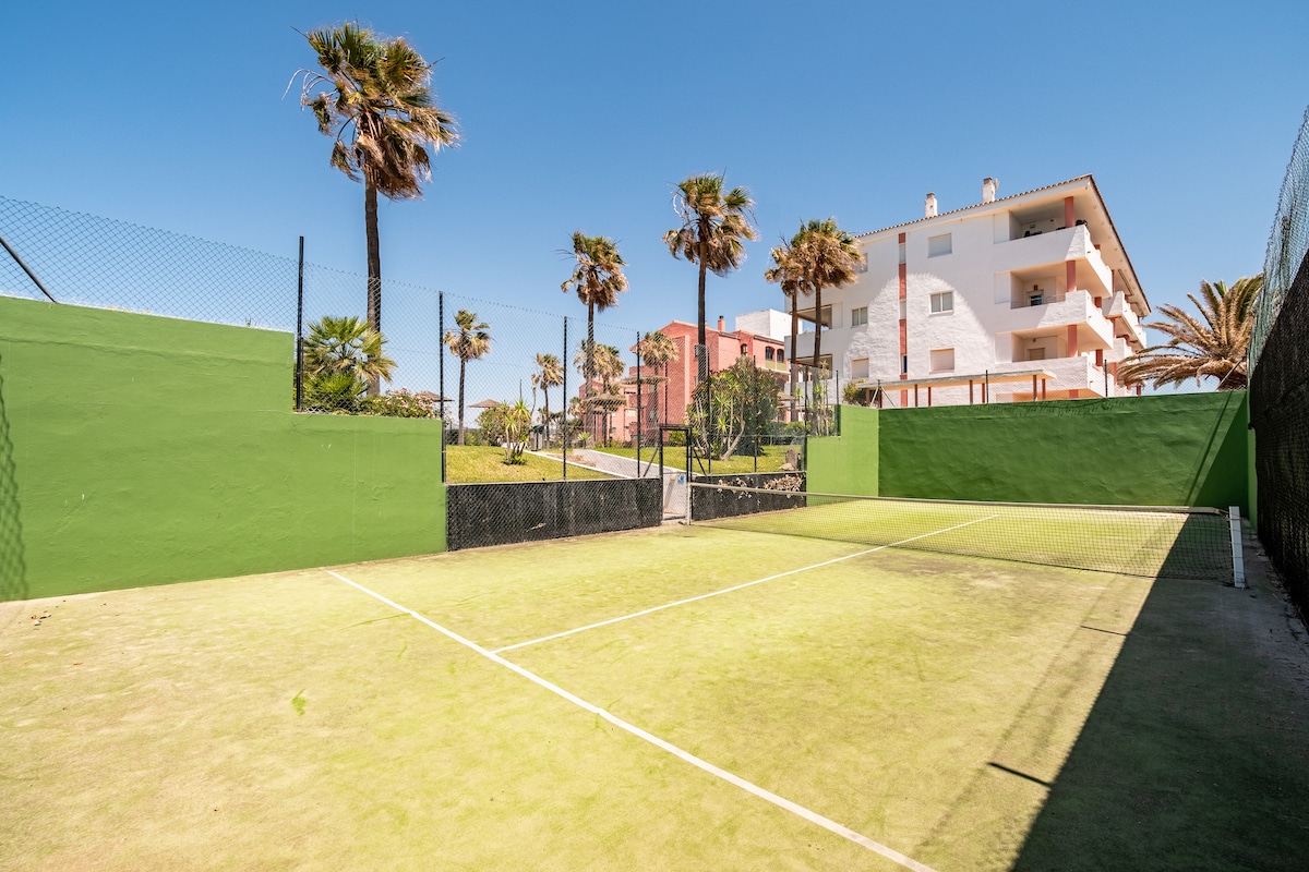 A tennis court is framed by palm trees and vibrant green walls, creating an open and spacious area for play. The court’s surface appears well-maintained, and a surrounding fence offers privacy. Nearby buildings can be seen in the background under a clear blue sky.