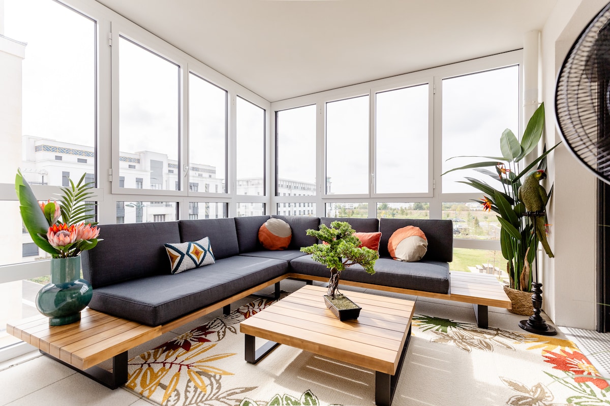 A sunlit living area features a spacious sectional sofa adorned with a decorative pillow, complemented by a wooden coffee table. Floor-to-ceiling windows provide ample natural light, while potted plants and vibrant flowers add a touch of nature, enhancing the tranquil atmosphere.