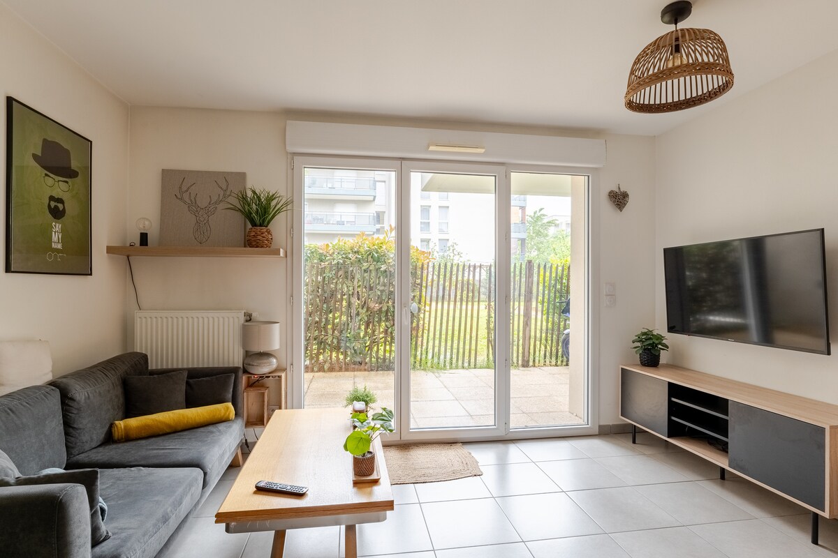 A living room is presented with a comfortable dark couch and a wooden coffee table. Large glass doors open to a private outdoor space, framed by greenery. A wall-mounted television complements a minimalist entertainment unit. Decorative elements include a plant on the table and wall art.