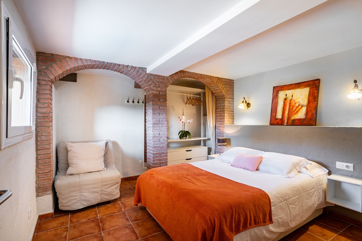 A cozy sleeping area features a spacious bed covered with an orange blanket. A chair is positioned in one corner, and a decorative wall art piece hangs nearby. Natural light enters through a window, illuminating the brick archways that enhance the room's charm.