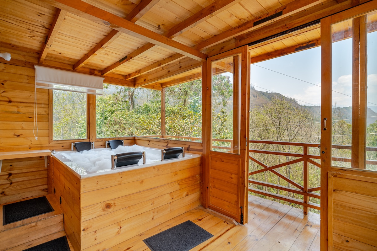 A wooden interior space is highlighted by a private jacuzzi, surrounded by large windows that offer views of the outdoor landscape. The room features a seating area with two chairs, enhancing the serene and natural atmosphere.