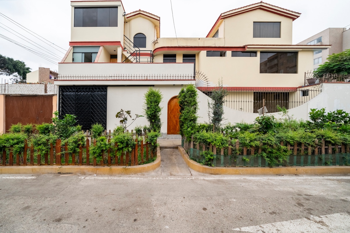 The exterior of a multi-level building showcases a blend of modern and traditional architecture. Lush greenery lines the path leading to a wooden door. Large windows provide natural light, while a private entrance is evident. A peaceful atmosphere is created by surrounding plants.