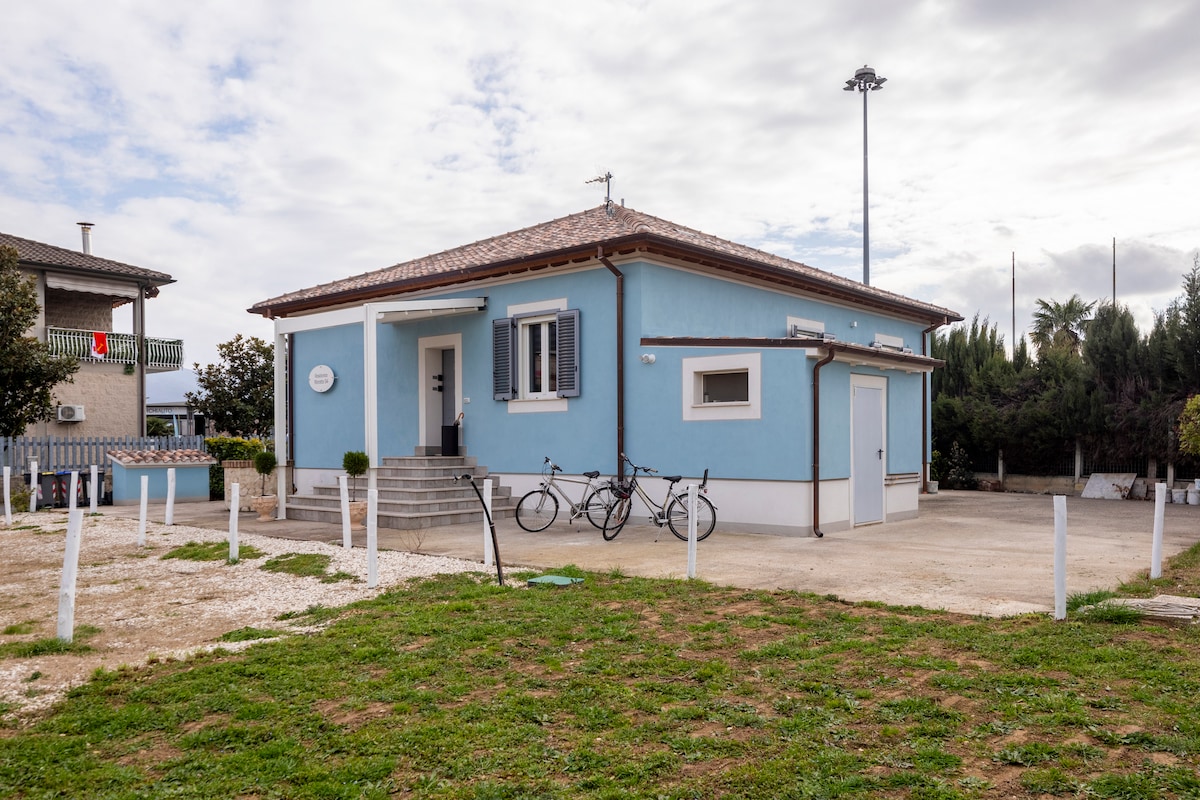 The exterior of the accommodation is presented, showcasing a light blue house with a sloped roof and white trim. A small entrance leads into the home, while bicycles are parked outside on a gravel surface. Lush greenery surrounds the property, creating an inviting space.