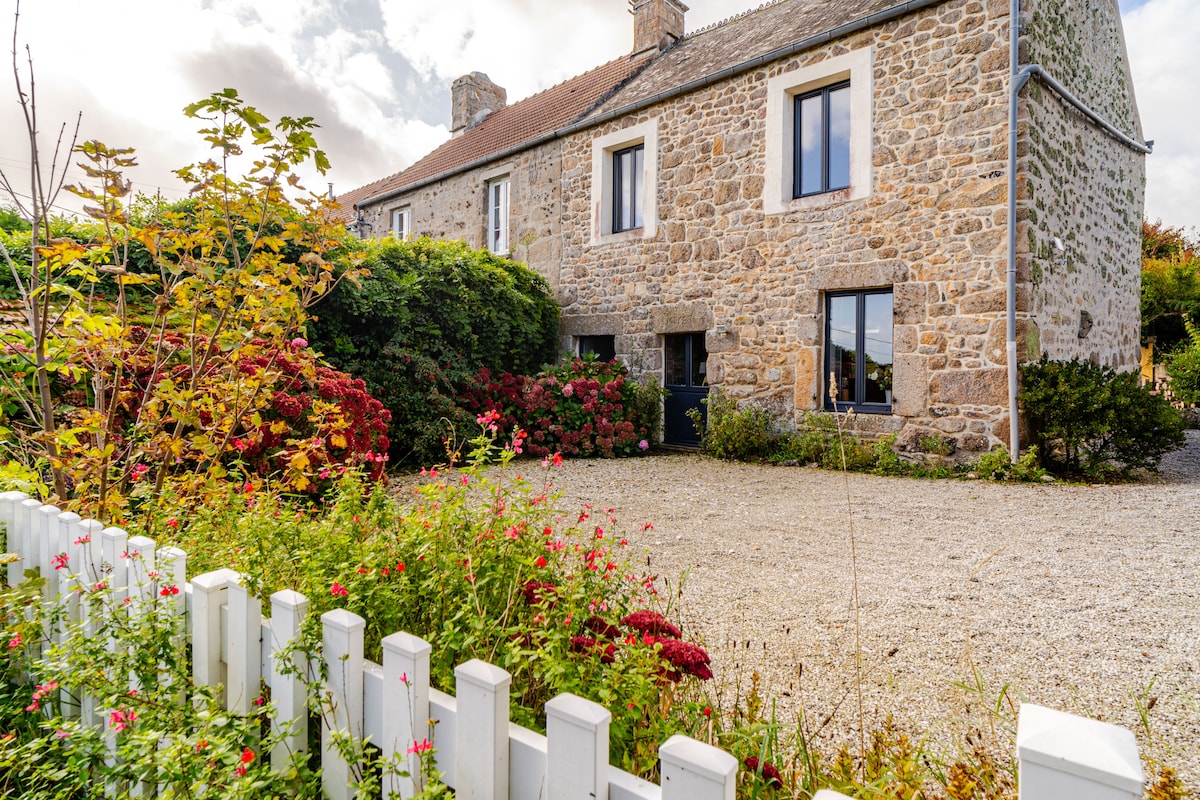 An authentic stone house is set amidst vibrant flowers and lush greenery. A gravel pathway leads to an entrance framed by flowering shrubs. Large windows reflect the natural light, creating an inviting exterior. A white picket fence outlines the property, enhancing its charm.
