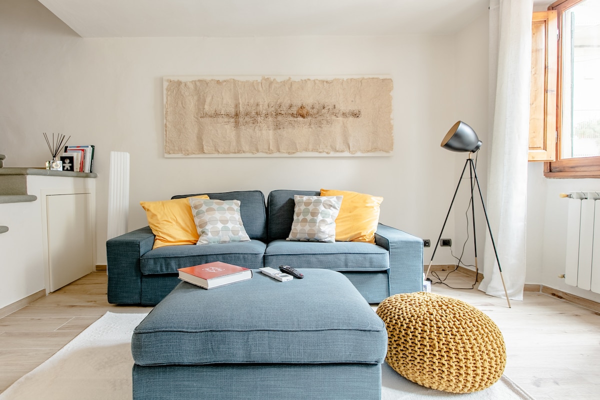 A bright living area features a comfortable blue sofa adorned with yellow cushions. A textured wall art piece is hung above the sofa, while a floor lamp stands beside it. Natural light enters through a nearby window, creating a welcoming ambiance.