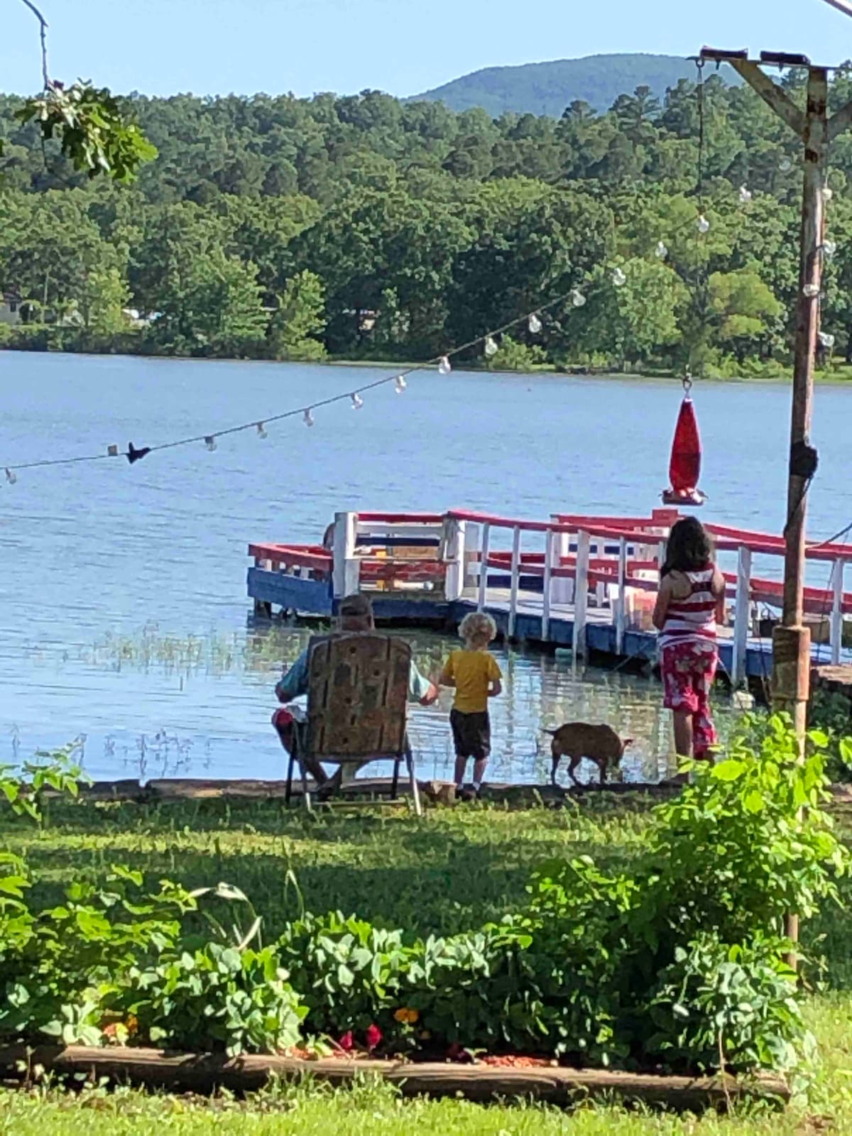 A serene lakeside view captures a family enjoying their time by the water. Two children and an adult are seen at the shore, while a cozy seating area with a person can be noted. Colorful plants frame the scene, enhancing the natural landscape.