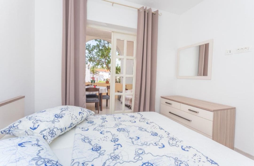A bright bedroom is depicted with a spacious bed adorned with a decorative blue and white patterned duvet. Natural light enters through a glass door leading to an outdoor space. A wooden dresser with a mirror and soft curtains frame the scene.