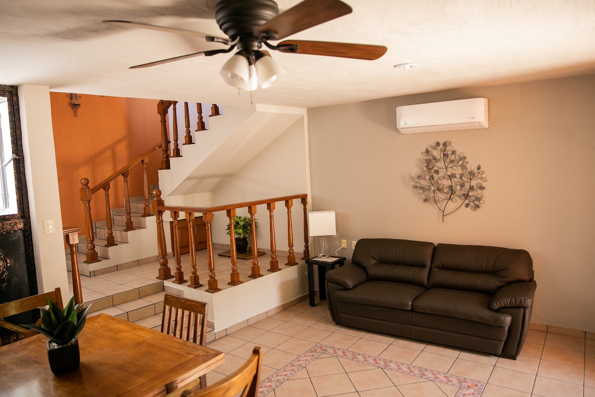 A spacious living area features a comfortable brown sofa positioned near a wooden dining table. A ceiling fan provides ventilation above. Light walls are complemented by a decorative wall piece, while a staircase leads to the upper level, creating an open and inviting space.