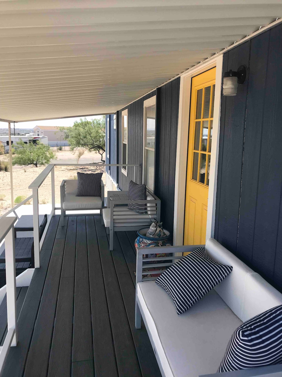 A covered porch features white sofas with navy striped cushions, positioned beside bright yellow doors. The flooring is a neutral-toned deck, creating a relaxed outdoor seating area. A small decorative item sits on a side table, adding to the welcoming ambiance of the space.