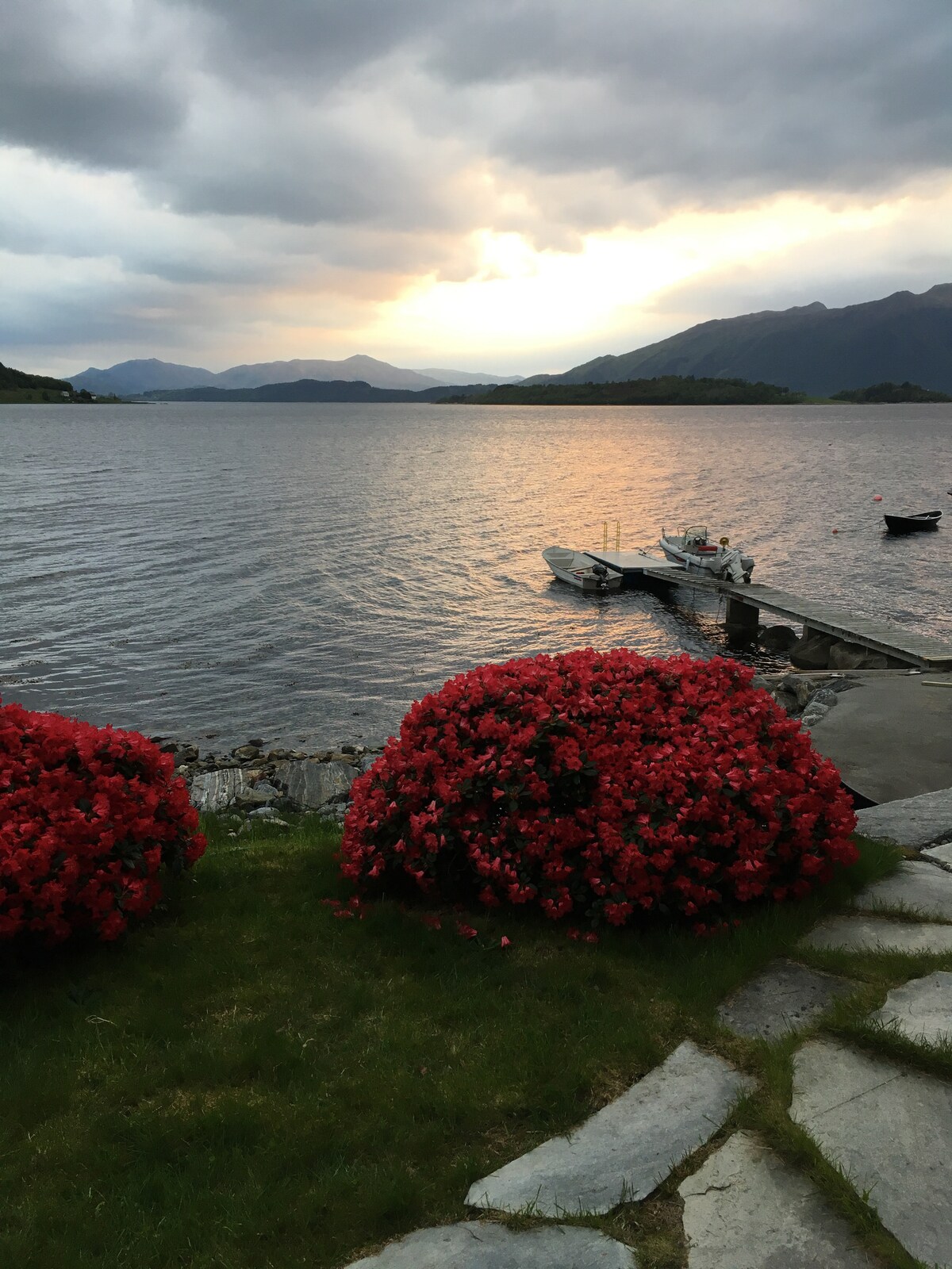 A view of calm waters reflects a muted sunset, framed by the distant mountains. Bright red flowering shrubs line a stone pathway leading to a wooden dock, where boats are moored. The cloudy sky adds to the serene atmosphere of the scene.