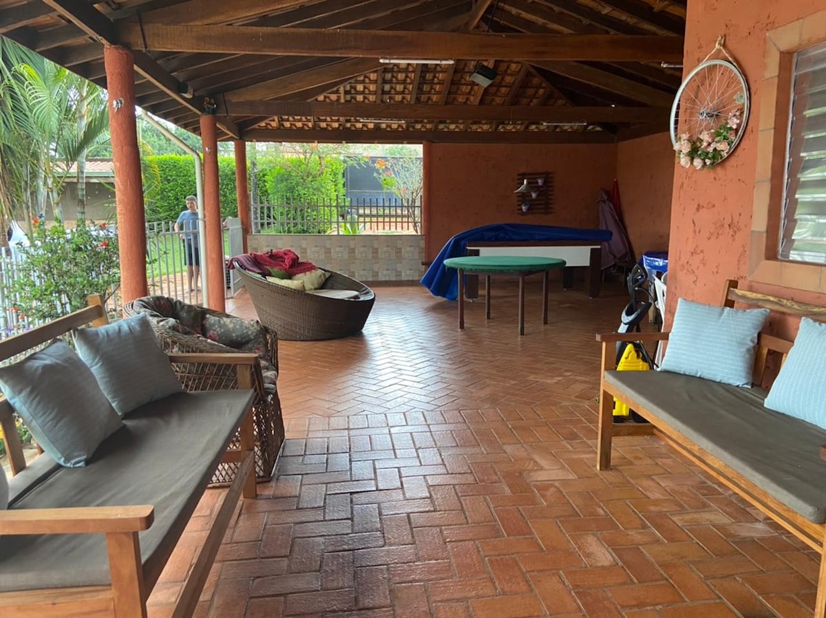 An open, covered area features a tiled floor and rustic wooden beams. Two wooden benches are positioned with soft cushions, alongside a round lounge chair. A pool table is accessible in the space, with greenery visible outside through the protective fencing.