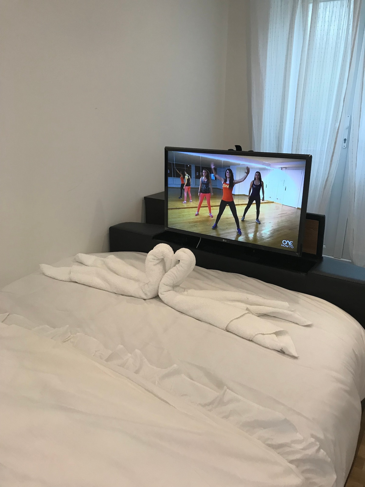 A spacious bed, neatly made with white linens, is positioned near a wall-mounted TV displaying a fitness program. Draped towels are arranged in the shape of swans on the bed, while sheer curtains allow natural light to filter into the inviting space.