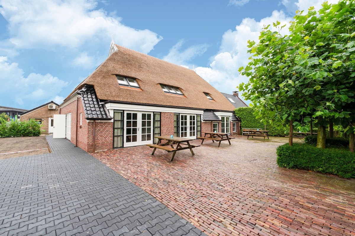 The exterior of the farm features a thatched roof and brick walls, surrounded by greenery. A spacious terrace with picnic tables is visible, set on a paved area. The entrance is highlighted, with large windows providing natural light, contributing to a welcoming outdoor environment.