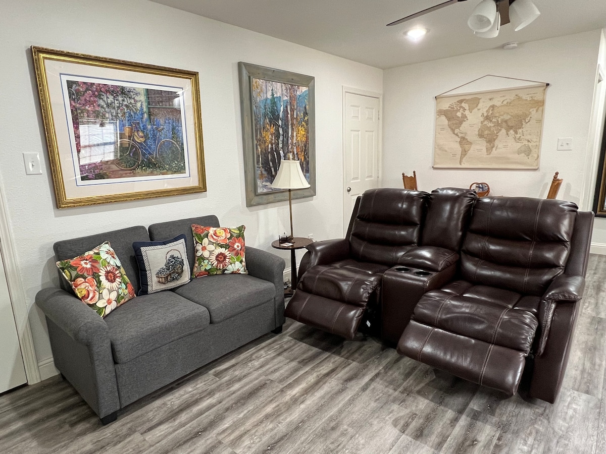 A living area features a gray sofa adorned with floral cushions and a recliner set in rich brown leather. A table lamp stands beside the recliners, while framed artwork and a world map hang on the wall, contributing to a well-decorated space.