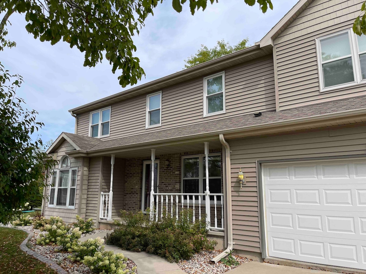 The exterior of the home is characterized by a welcoming front porch with a white railing, surrounded by landscaped flower beds. A garage door is visible on the right, and multiple windows adorn the front facade, allowing natural light into the living space.