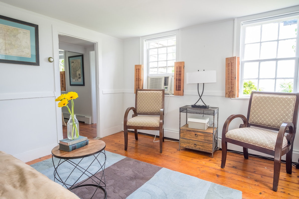 The living area features two patterned armchairs positioned across from a circular coffee table. A side table holds a lamp, and a vibrant bouquet of yellow flowers adds a cheerful touch. Natural light fills the room through two windows, enhancing the welcoming environment.