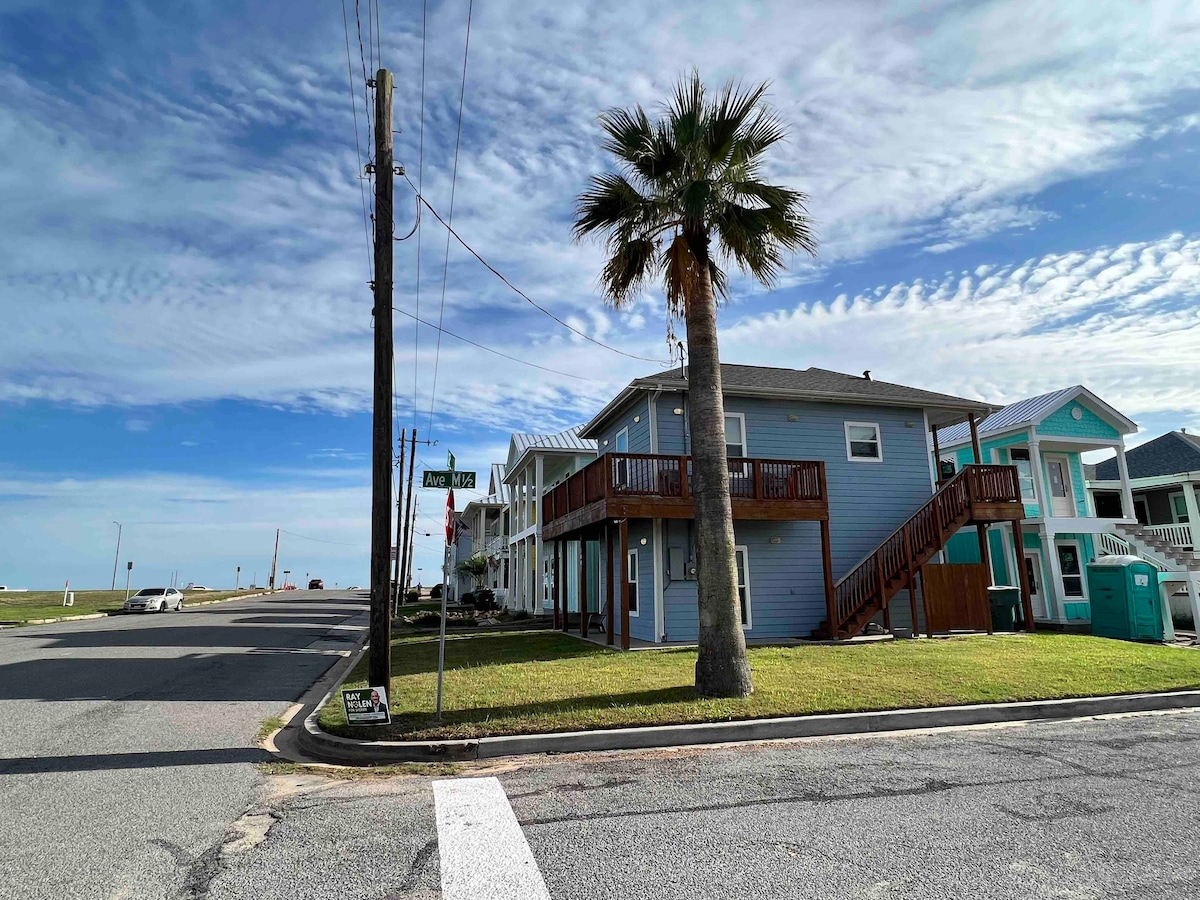 A corner lot features a light blue two-story house with a balcony and a staircase. Nearby, palm trees and grassy areas are visible. The street sign indicates 'Sandy Lane' while the beach is seen in the distance, framed by a clear blue sky with scattered clouds.
