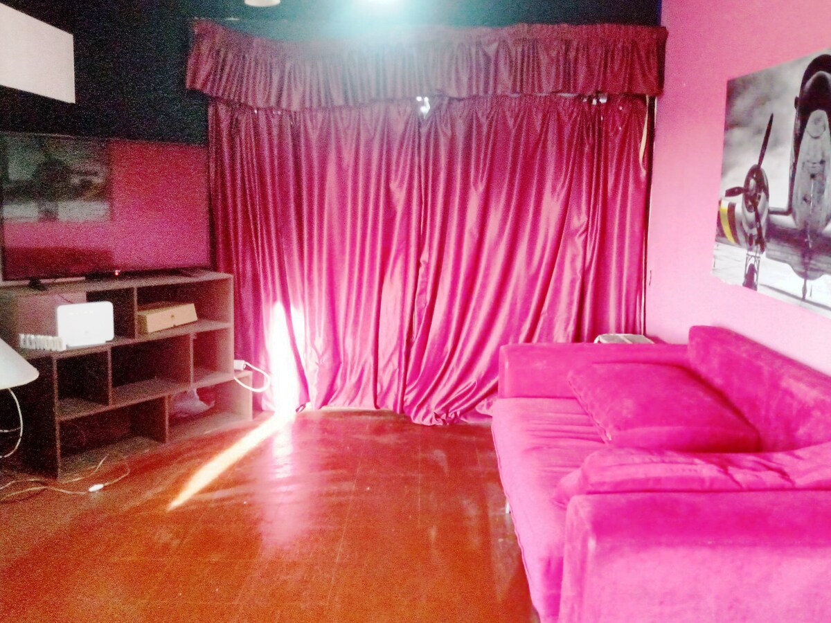 A cozy living area features a plush pink sofa set against a deep blue wall. A large television is mounted on the opposite wall, while floor-to-ceiling curtains in rich burgundy provide a dramatic touch. Natural light filters in, highlighting the warm wood flooring.