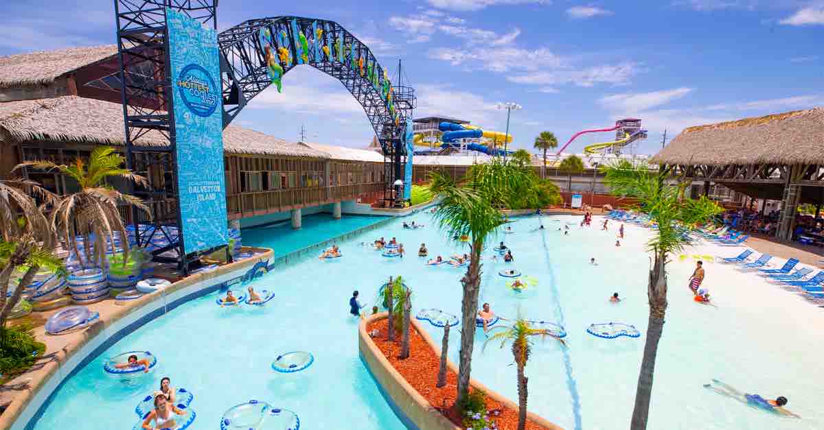 A vibrant water park scene displays a large wave pool surrounded by palm trees. Colorful water slides tower above. Guests float on inner tubes while others enjoy the water's edge. Thatched-roof structures provide shaded seating areas, enhancing the lively atmosphere for all ages.