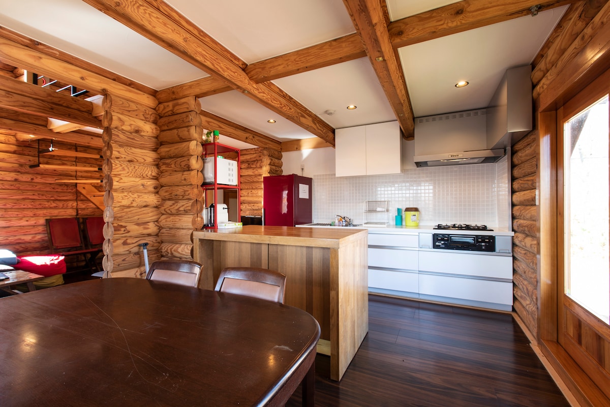 A well-equipped kitchen area features wooden beams and cabinetry. A central island provides ample workspace, surrounded by a dining table with four chairs. Modern appliances, including a refrigerator and gas stovetop, are visible, with natural light streaming in from a nearby window.