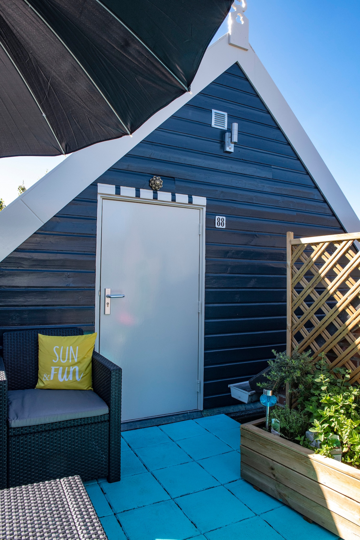 The exterior of the studio is shown, featuring a sloped roof and a white door marked with the number 38. A cozy seating area with a cushion reading 'SUN' is visible, under a large umbrella, alongside a small planted garden and lattice fencing.