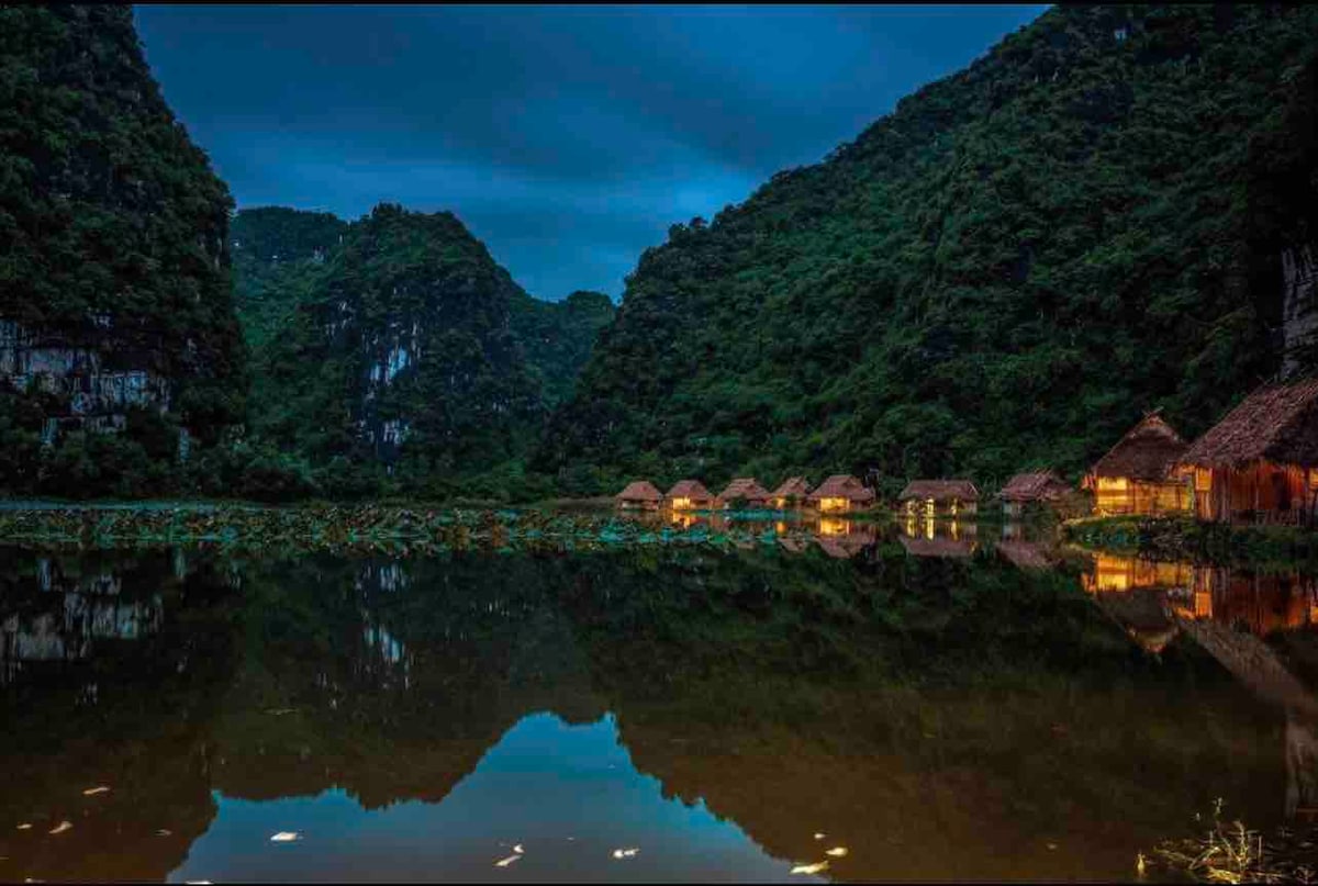 A serene lake is bordered by lush mountains, reflecting charming bungalows with thatched roofs. The gentle lights from the bungalows illuminate the surrounding water, creating a peaceful atmosphere. The deep greens and blues of the landscape complement the tranquil scene.